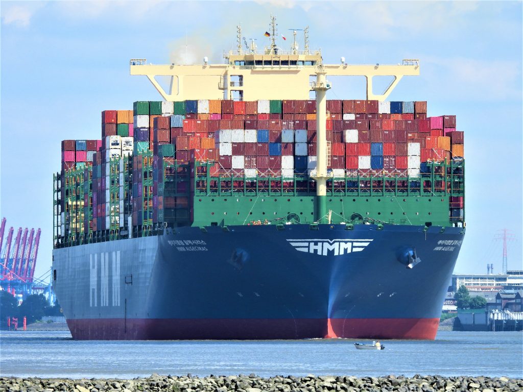 Großes Containerschiff in einem Hafen, im Vordergrund ein kleines Motorboot, in dem eine Person sitzt.