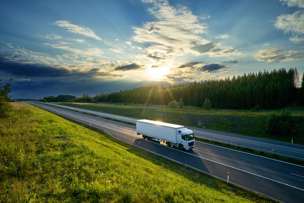 Ein weißer LKW fährt auf der Autobahn durch die Landschaft im Licht der untergehenden Sonne mit dramatischen Wolken.